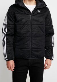 Svart quiltad jacka med dragkedja och huva. Har vita tre-randiga detaljer på ärmarna och en diskret logotypmönster över hela.