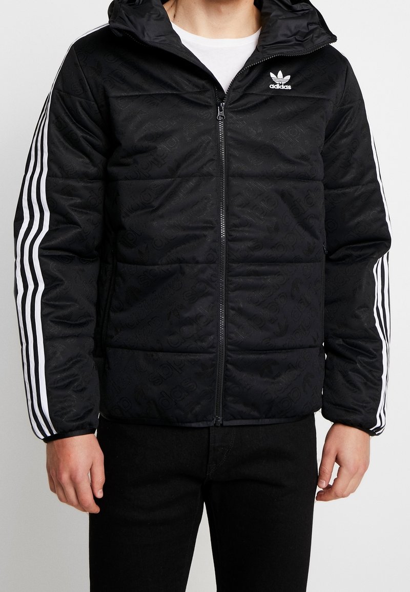 Svart quiltad jacka med dragkedja och huva. Har vita tre-randiga detaljer på ärmarna och en diskret logotypmönster över hela.