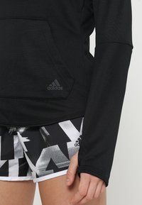 Primo piano di una persona che indossa un hoodie nero Adidas e pantaloni corti a motivi bianco e nero Adidas, con la mano rilassata al suo fianco.