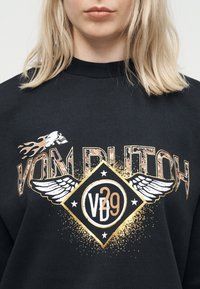 Γυναίκα που φοράει μαύρο φούτερ με το κείμενο «Von Dutch», γραφικό με φλογερό κρανίο και έμβλημα με φτερά που αναγράφει «VD 29» μπροστά.