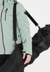 Vihreä eristävä takki, jossa on mustat vetoketjut, varustettu mustalla laukulla, jossa on kantohihnat ja logo. Rakenteellinen kangas ja toiminnallinen muotoilu.