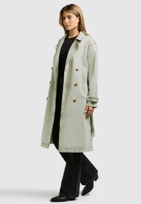 Donna con cappotto trench lungo verde chiaro sopra camicia nera e pantaloni neri, in piedi davanti a uno sfondo uniforme grigio chiaro.