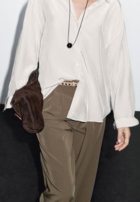 White button-up shirt with loose fit, paired with olive green trousers, holding a dark brown suede bag, featuring a tan belt with round holes.