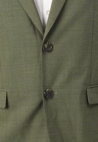 Blazer verde oliva con un tejido texturizado, dos botones oscuros y un bolsillo en la solapa del pecho. El diseño está ajustado con un clásico cuello.