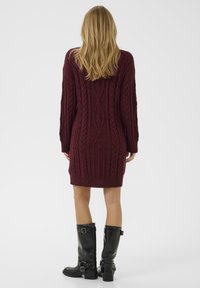 Burgundska pletena obleka s teksturiranimi kabli, dolge rokave in oprijet dizajn, kombinirana s črnimi škornji do kolen z zaponkami.