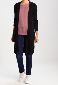 Cardigan noir en maille avec ouverture frontale et poches, porté sur un haut texturé rose clair et un pantalon bleu foncé, accompagné de baskets blanches.