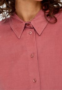 Roze corduroy shirt met een puntige kraag, voorzien van een verticale ribbelstructuur en vijf knopen. De stof heeft een zachte uitstraling en afwerking.
