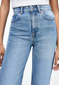 Mujer con jeans de denim de tiro alto, de color azul claro, y una blusa sin mangas de textura blanca, de pie con las manos relajadas a los lados.