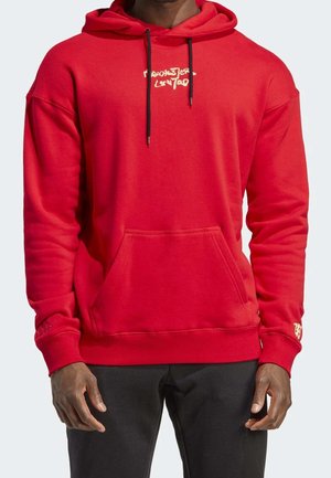 Homme portant un sweat à capuche rouge avec une poche avant et des cordons noirs, associé à un pantalon noir, debout devant un fond uni.