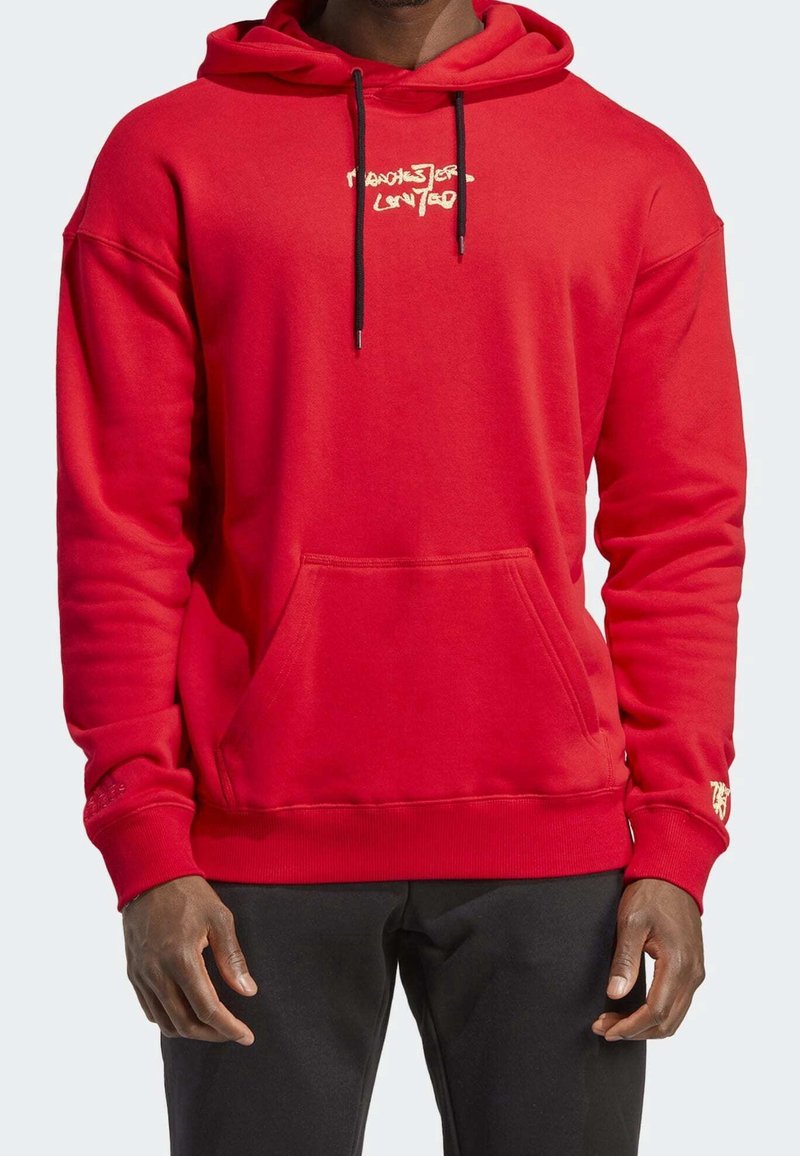 Homme portant un sweat à capuche rouge avec une poche avant et des cordons noirs, associé à un pantalon noir, debout devant un fond uni.