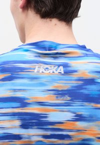 Πίσω όψη ενός ατόμου που φοράει αθλητικό μπλουζάκι HOKA με μοτίβα σε μπλε, πορτοκαλί και άσπρο χρώμα, με κοντά καστανά μαλλιά ορατά.