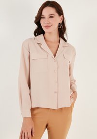 Svjetlopinkasta bluza s kragnom, dugim rukavima, dva prednja džepa i kopčanjem na gumb. Izrađena od glatke tkanine, karakterizira opušteni kroj.
