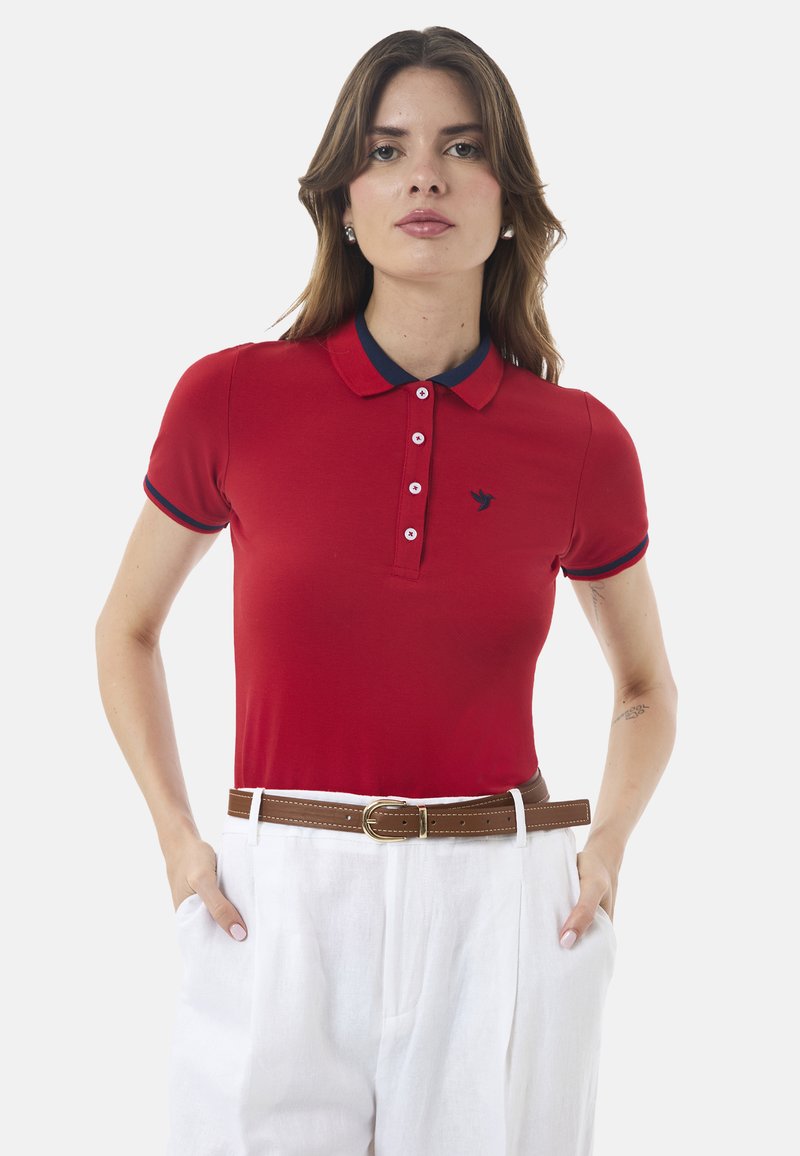 Rød polo skjorte med en marineblå krave, knaplukning og broderet logo. Kombineret med hvide linnedbukser og et brunt bælte.