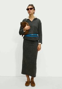 Ciemnoszary sweter z dzianiny z białym kołnierzykiem, dopasowana długa spódnica, niebieski pasek i brązowe buty. Modelka trzyma brązową torbę, nosi okulary przeciwsłoneczne.