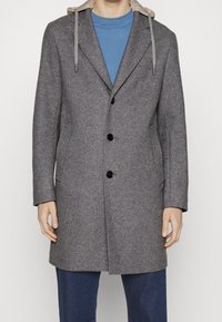 Uomo che indossa un cappotto grigio con bottoni e cappuccio beige, abbinato a una camicia blu e pantaloni blu scuri, in piedi contro uno sfondo neutro.