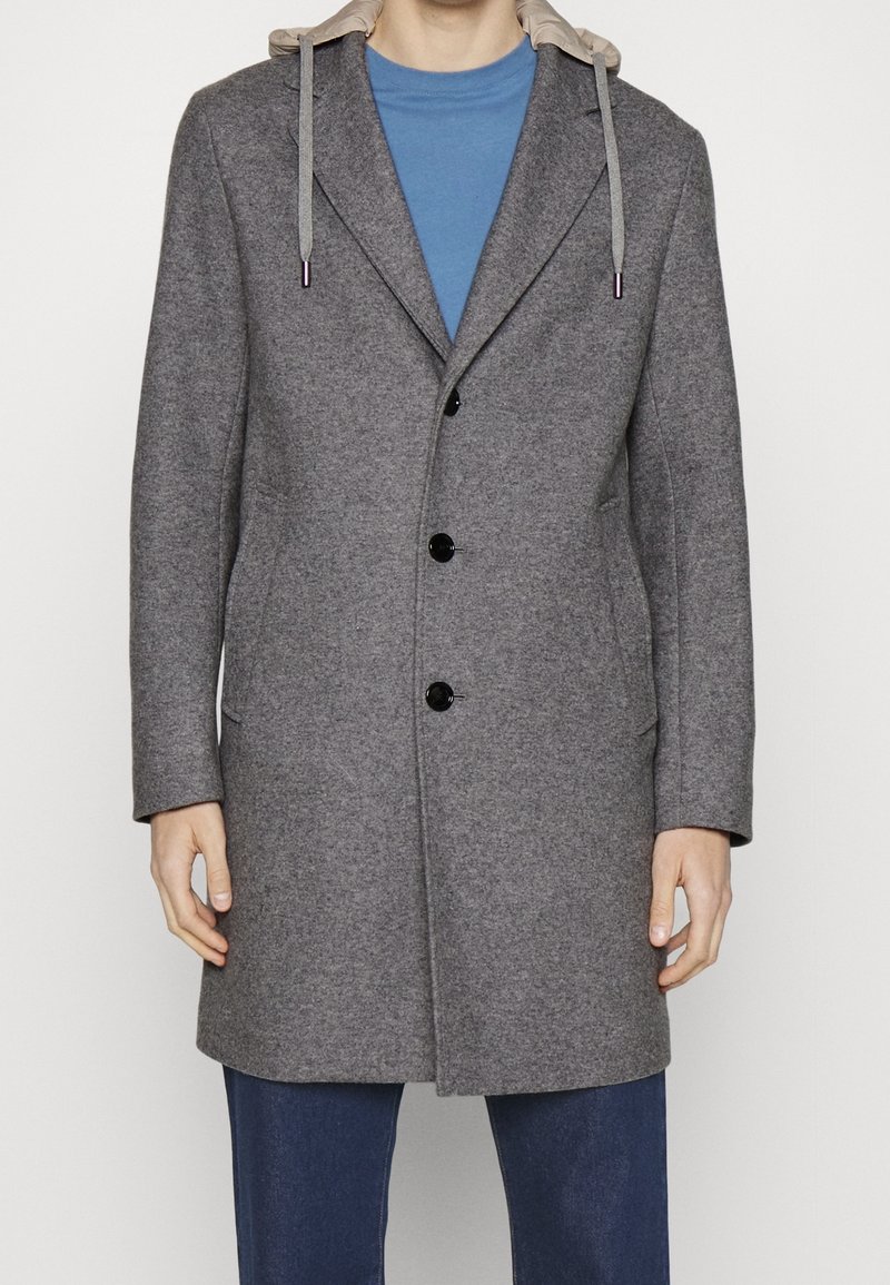 Uomo che indossa un cappotto grigio con bottoni e cappuccio beige, abbinato a una camicia blu e pantaloni blu scuri, in piedi contro uno sfondo neutro.