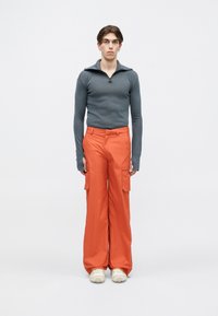 Grijze lange mouwen top met ritsnek; relax-fit oranje cargo broek; crèmekleurige chunky sneakers; effen achtergrond.