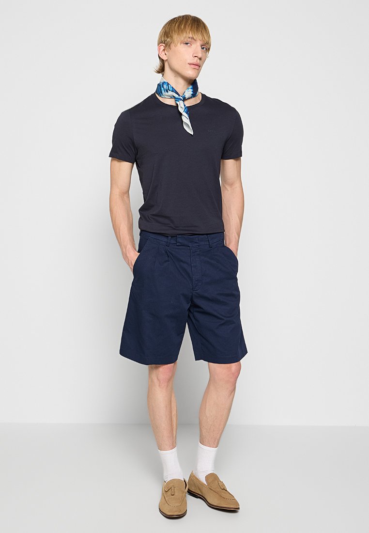 Marineblaue Shorts aus Baumwollmischung, kombiniert mit einem eng anliegenden schwarzen T-Shirt. Accessories sind ein hellblau-weiß gemusterter Halstuch und beige Schuhe.