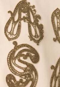 Broderie de perles dorées ornées formant des lettres stylisées sur un tissu crème, mettant en valeur un savoir-faire détaillé et un motif décoratif.