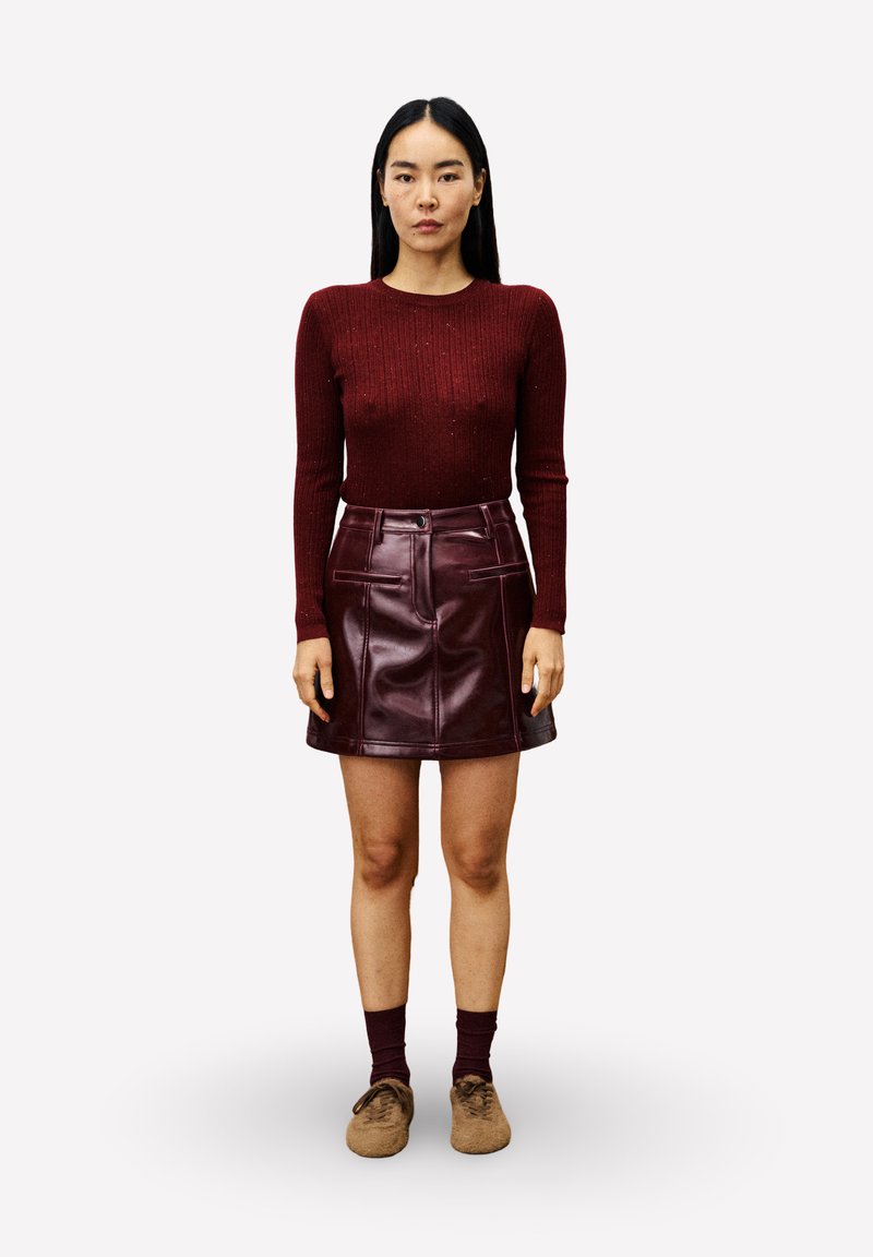 Vrouw staand, naar voren gericht, gekleed in een bordeauxrode geribbelde trui en een glanzende bordeauxrode leren rok met bruine schoenen en bijpassende sokken.
