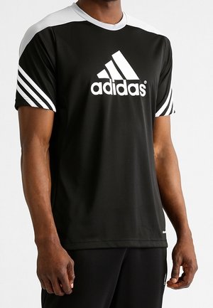 Mężczyzna w czarnej sportowej koszulce Adidas z białymi panelami na ramionach i trzema białymi paskami na rękawach, stojący na jednolitym tle.
