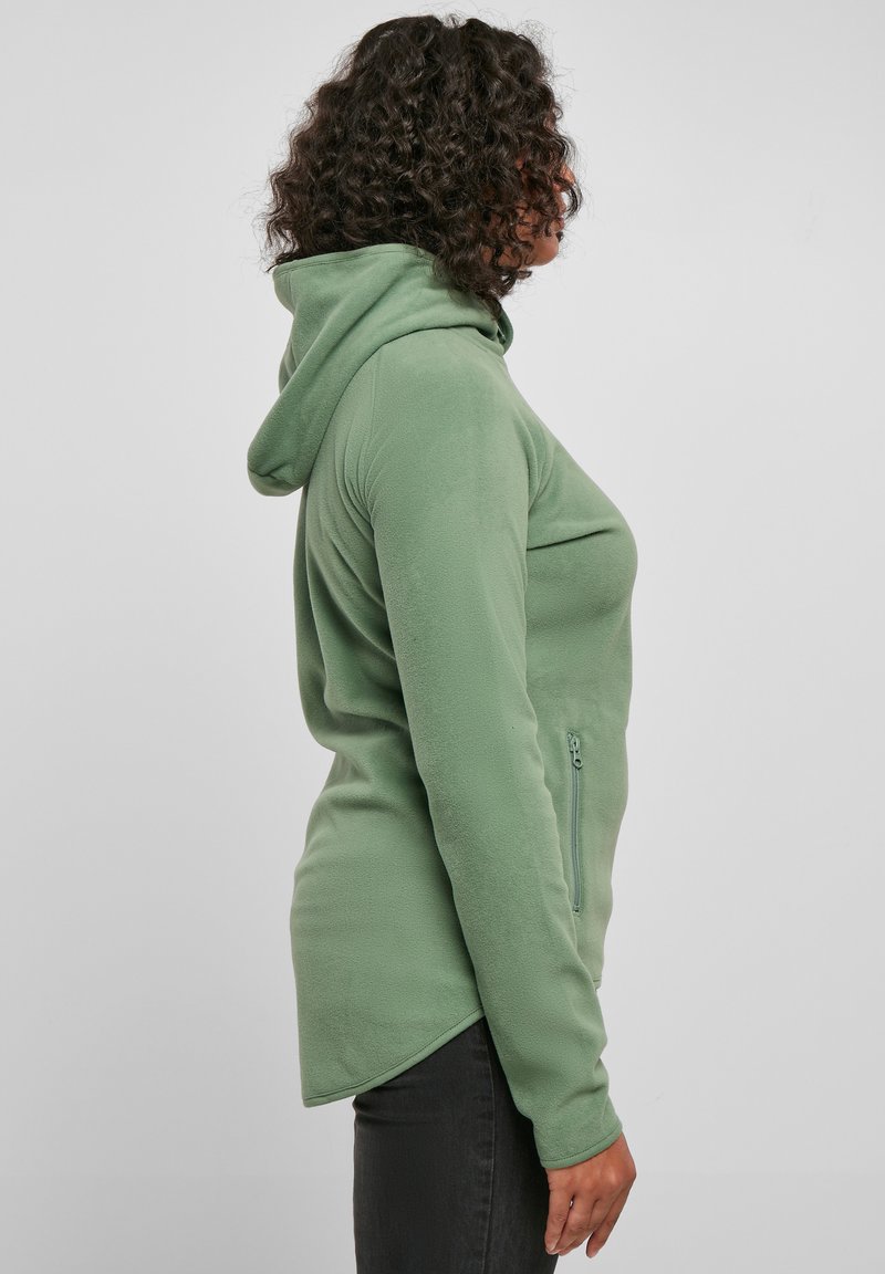 Felpa verde in pile con cappuccio, tasca laterale con zip, orlo arrotondato e maniche lunghe. Texture liscia e design casual, silhouette attillata.