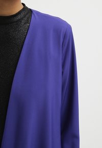 Lila jacka med öppen front i mjukt tyg, med långa ärmar och avslappnad passform, lager-på-lager över en svart glansig topp.