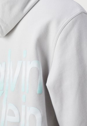 Gros plan d'un sweat à capuche gris clair montrant un logo "Calvin Klein" bleu pâle sur un tissu doux près de la couture de l'épaule et de la capuche.