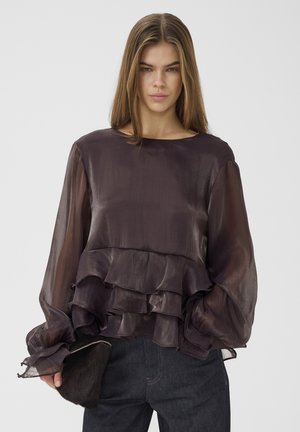 Femme portant un chemisier violet foncé transparent avec des volants superposés et des manches bouffantes, tenant une pochette noire texturée sur un fond uni.