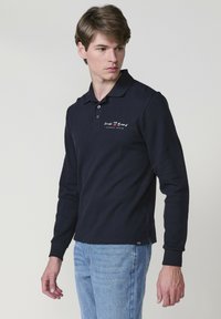 Tengerészkék, hosszú ujjú pólóing, texturált anyagból készült, felgombolható gallérral és a bal mellen varrott logóval.