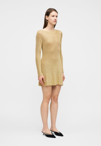 Γυναίκα που φοράει ένα μακρυμάνικο χρυσό πλεκτό μίνι φόρεμα και μαύρες μπαλαρίνες με λεπτό τακούνι και ιμάντες, στέκεται μπροστά σε ένα απλό λευκό φόντο.