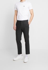 Mężczyzna ubrany w białą koszulkę Calvin Klein, ciemnoszare dopasowane spodnie, białe skarpetki i białe sneakersy, stojący na tle czystej białej ściany.