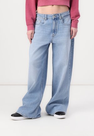 Person iført lyseblå vidde bukser, sorte og hvide sneakers og en kort pink langærmet top, stående mod en hvid baggrund.