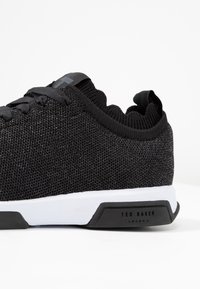 Schwarze Strick-Sneaker mit strukturiertem Obermaterial, gewelltem Kragen und einer weißen Zwischensohle. Enthält ein markiertes schwarzes Gummi-Detail an der Ferse.