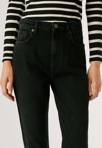 Augstas jostas melni denimi džinsi ar strukturētu apdari, ar zelta tonētiem detaļām un sānu kabatām, kombinēti ar melnbaltu svītrainu topu.