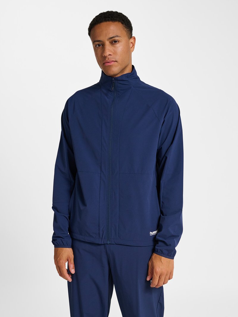 Ein Mann steht und trägt eine marineblaue Jacke mit Reißverschluss und passende Hose vor einem schlichten weißen Hintergrund.