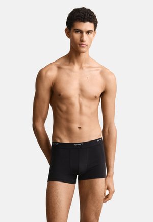 Junger Mann steht in schwarzen GANT-Boxershorts und blickt nach vorne vor einem einfarbigen hellen Hintergrund.