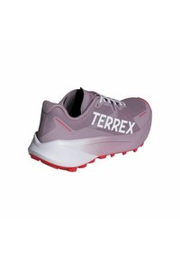 Scarpa da corsa viola chiaro con logo "TERREX" bianco, caratterizzata da una tomaia testurizzata, intersuola bianca e suola in gomma rossa con lugs di trazione pronunciati.