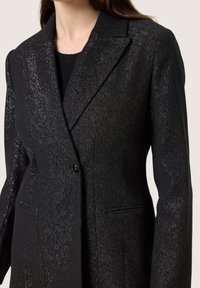 Közelkép egy illeszkedő fekete zakóról, finom ezüst csillogással, hajtókás gallérral és egysoros gombolással, fekete top felett.