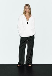 Vit oversized blus med djup V-ringning, kombinerad med lösa svarta byxor. Setet fulländas med ett svart hängehalsband.