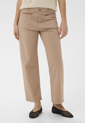 Kvinde iført beige bukser med lige ben og store forlommer samt sorte vævede flade sko, stående mod en ensartet baggrund.