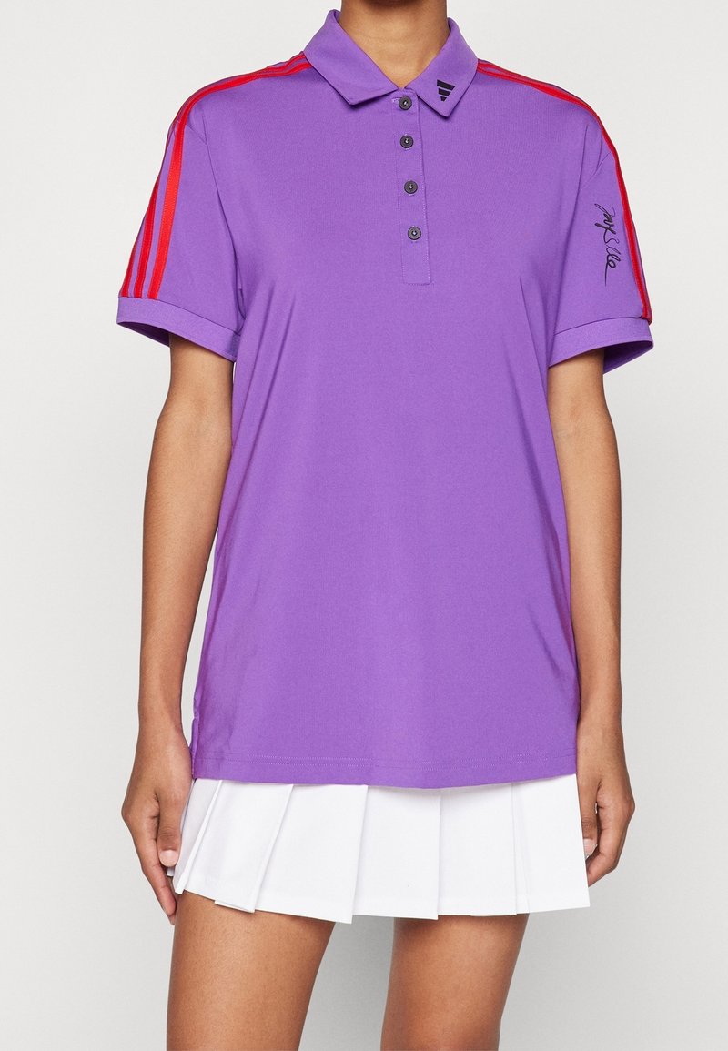Personne portant un polo violet avec des rayures rouges sur les manches et une jupe plissée blanche.