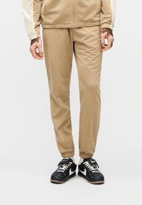 Χακί φόρμες με ελαστικά μανσέτες, με λεία υφή και μινιμαλιστικό σχεδιασμό. Φοριούνται με μαύρα αθλητικά παπούτσια και μπεζ μπουφάν.