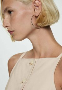 Krémový bezrukávový top se rovnou výstřihem, se zdobnými knoflíky. Modelka má krátké blond vlasy a nosí velké stříbrné naušnice ve tvaru kruhu.