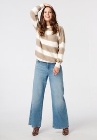 Beige en wit gestreepte gebreide trui, wijde lichtblauwe spijkerbroek en dierenprint hoge hakken; model staat met één hand op het hoofd.