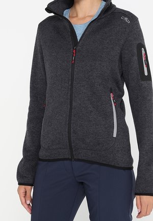 Femme portant une veste polaire zippée gris foncé avec des fermetures éclair aux accents rouges et une chemise bleu clair en dessous, associée à un pantalon bleu marine.