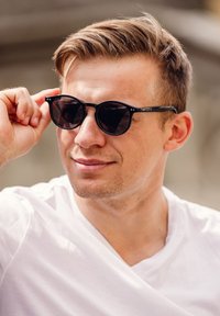 Černé kulaté sluneční brýle s tmavými skly, ukazující tenký rám. Model má krátké vlasy a na sobě má bílé tričko.