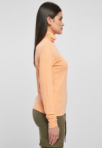 Maglia a maniche lunghe color pesco con collo alto realizzata in tessuto leggero. Caratterizzata da una silhouette aderente e una texture morbida. Indossata con pantaloni verde oliva.