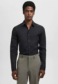 Cămașă neagră cu mânecă lungă, cu un design ajustat, textură netedă și închidere cu nasturi, asortată cu pantaloni gri deschis.