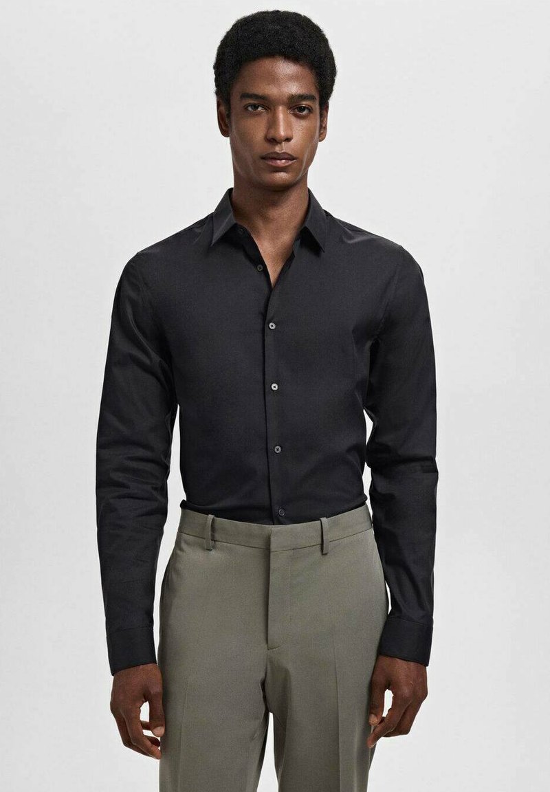 Cămașă neagră cu mânecă lungă, cu un design ajustat, textură netedă și închidere cu nasturi, asortată cu pantaloni gri deschis.